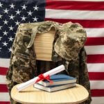 Diploma and books on a circular table and military uniform on a wooden chair and a US flag behind them