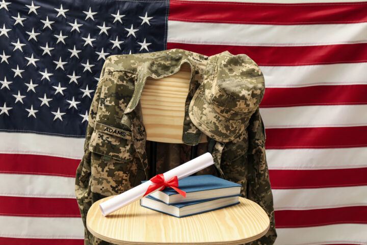 Diploma and books on a circular table and military uniform on a wooden chair and a US flag behind them