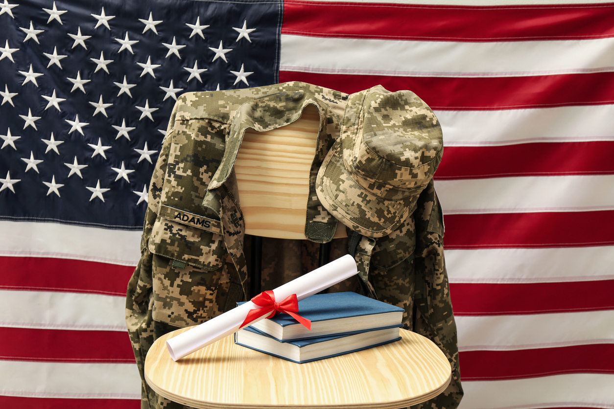 Diploma and books on a circular table and military uniform on a wooden chair and a US flag behind them